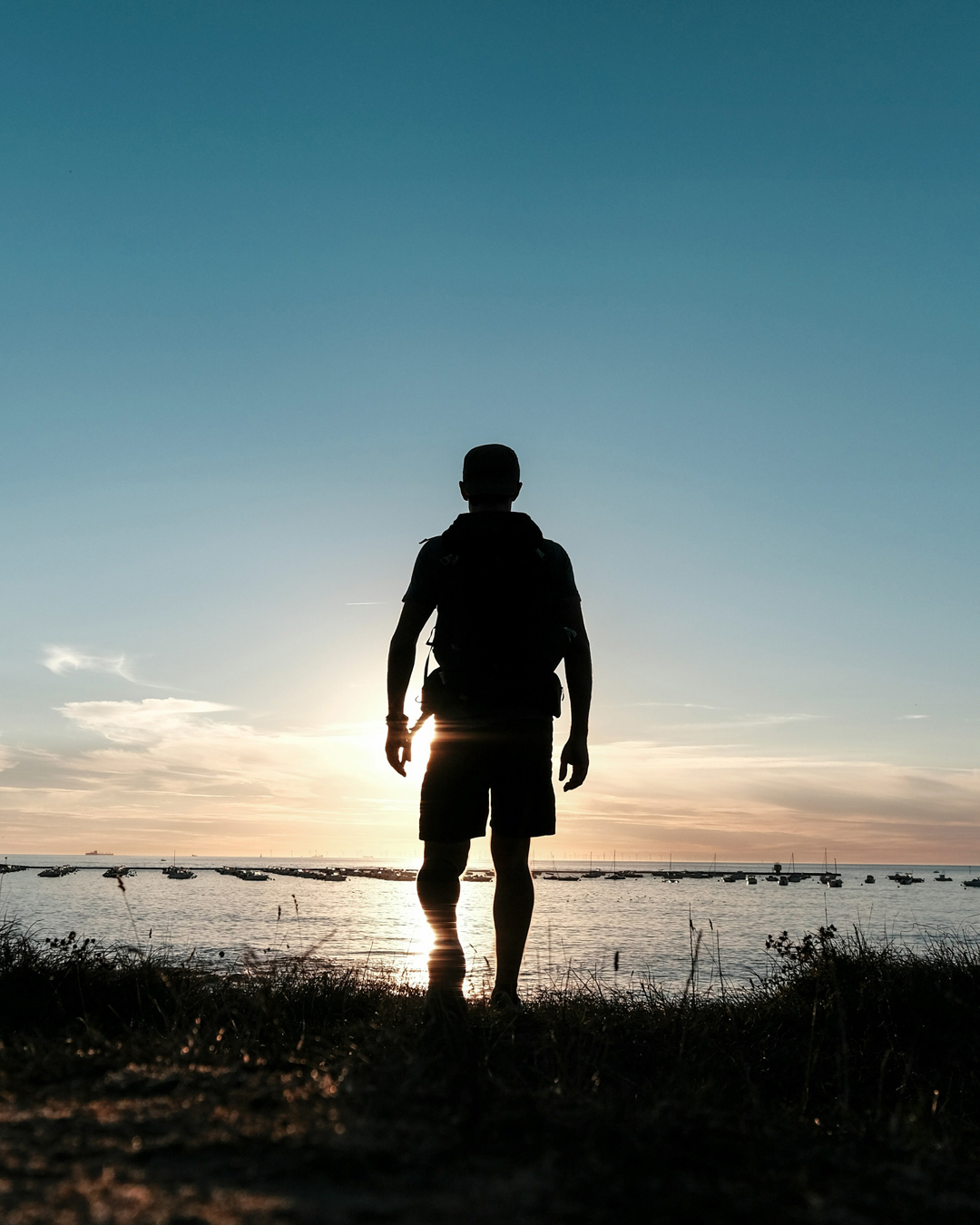 Silueta de una persona con una mochila de pie en la orilla al atardecer viendo el horizonte.